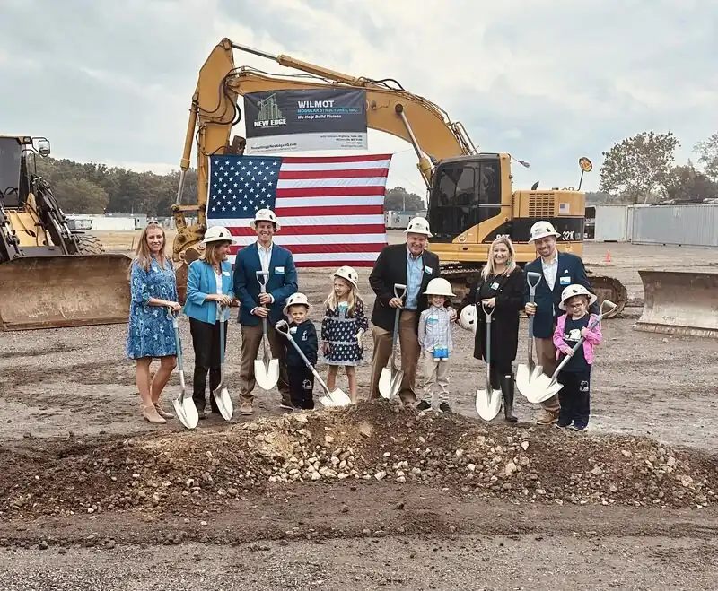 The Wilmot family in hard hats with ceremonial shovels at a groundbreaking, in front of an excavator, a Wilmot banner and an American flag