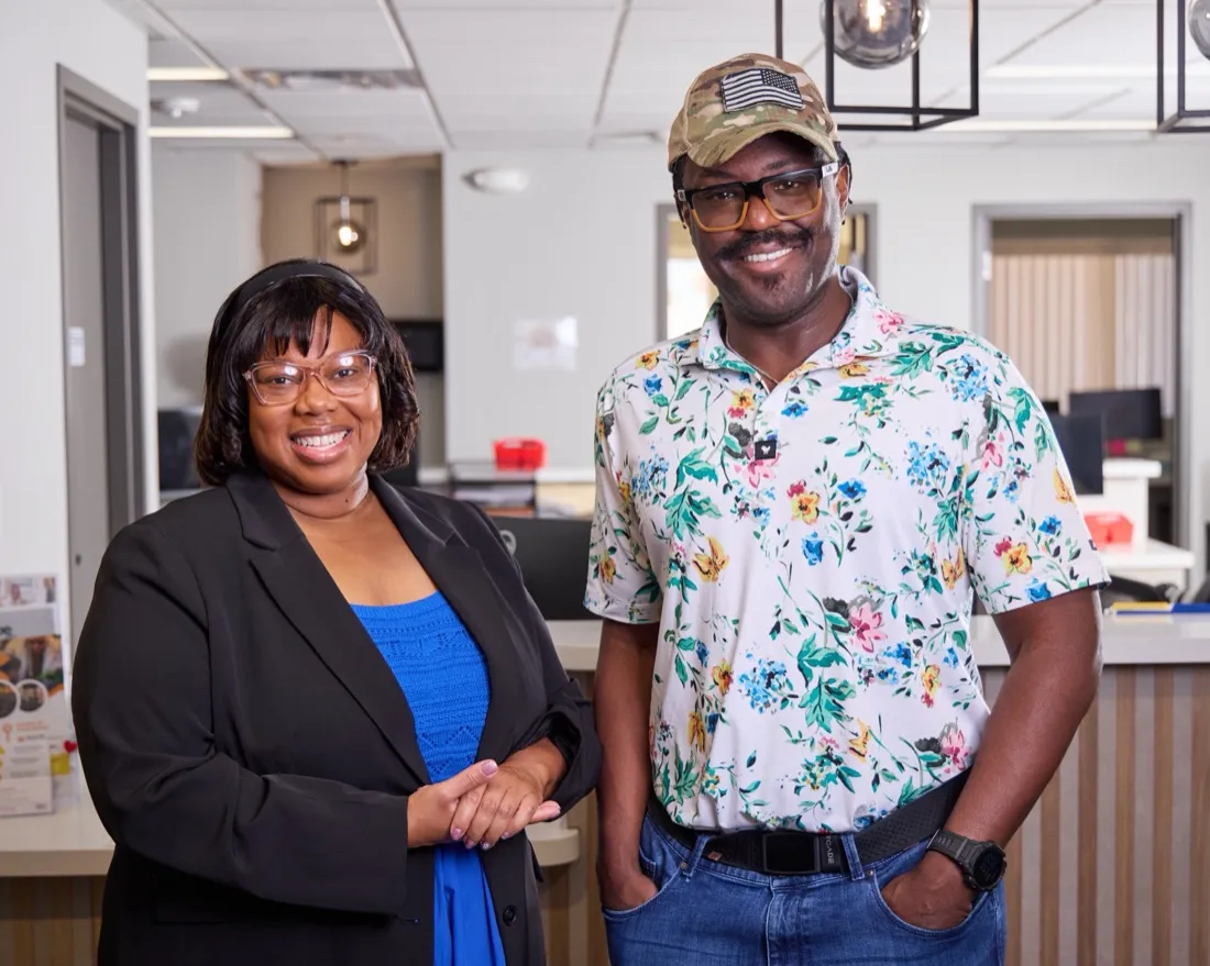 Kamilah Bywaters in a black blazer standing next to a smiling man in a floral shirt and camouflage hat