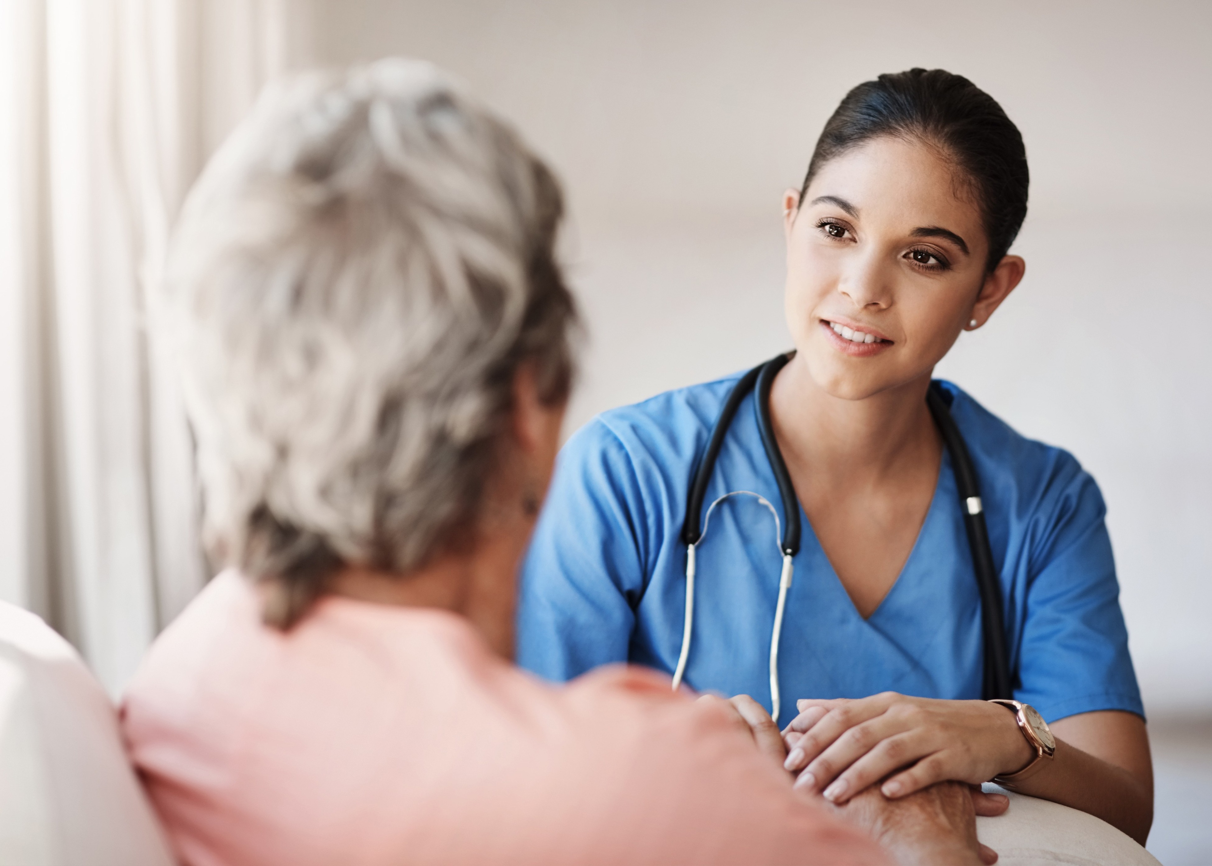 Young nurse in blue scrubs attentively holding the hands of an elderly patient.