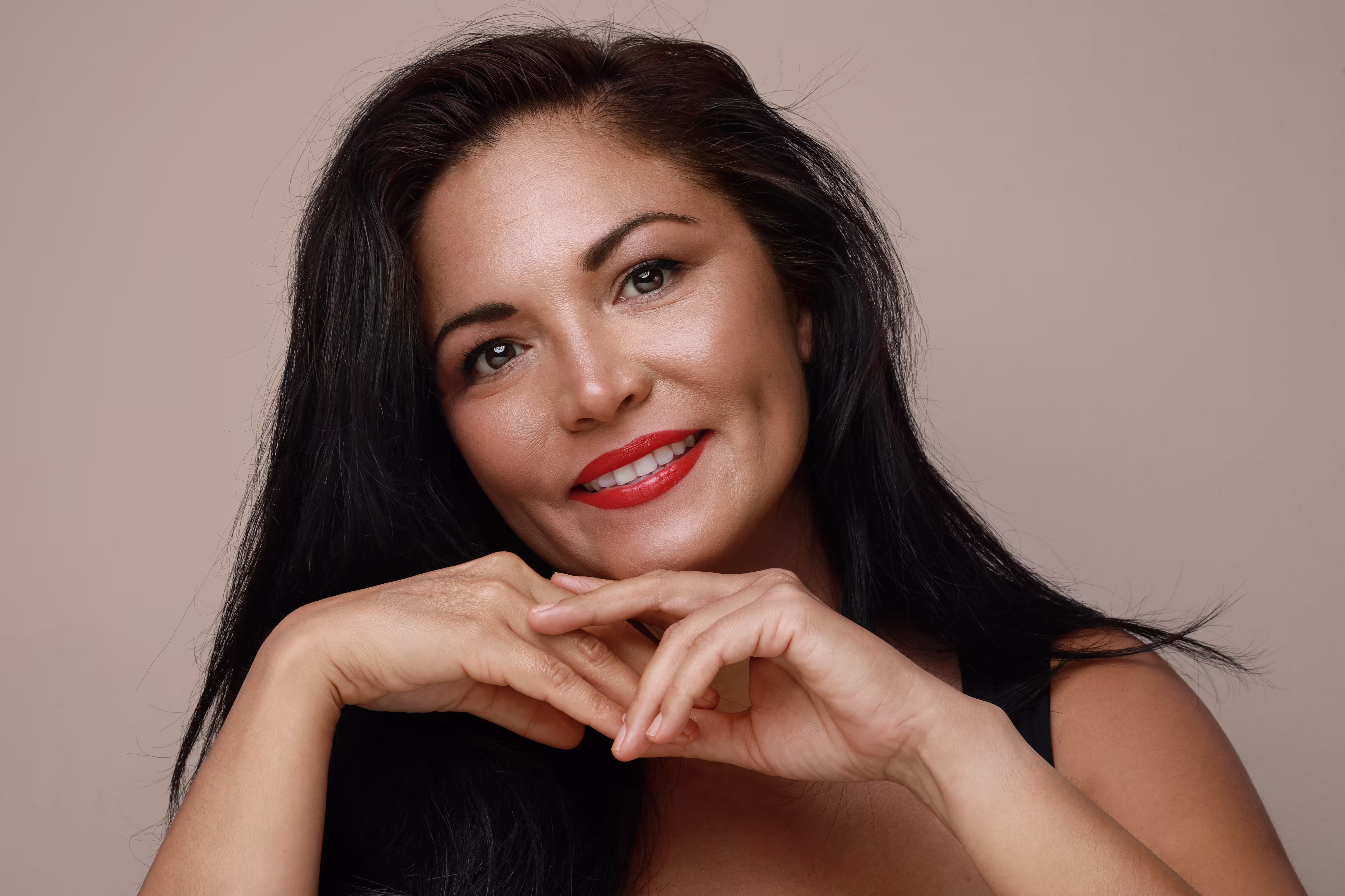 Smiling woman with long black hair, red lipstick, and hands gently touching under her chin against a beige background.
