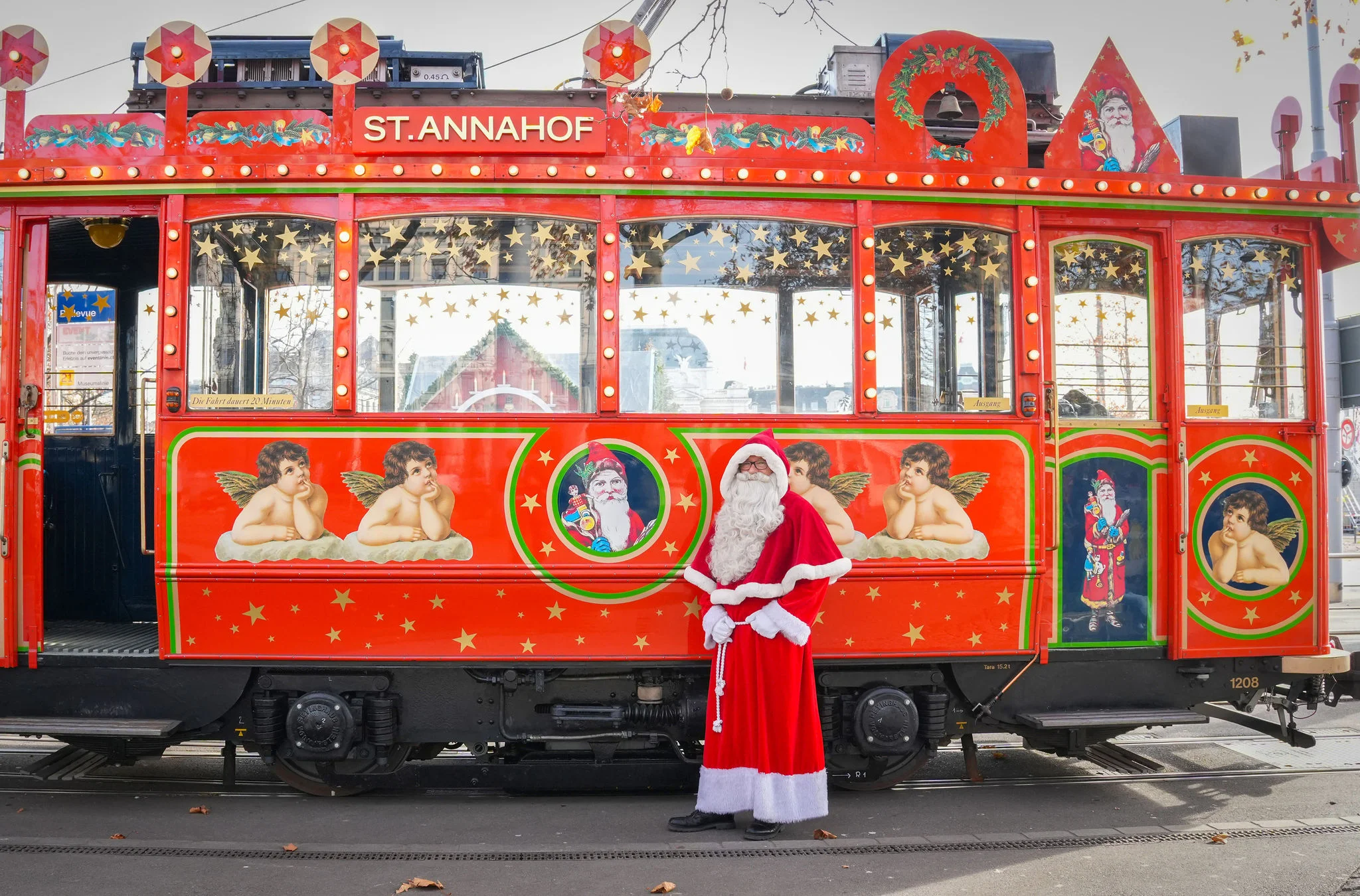 Weihnachtlich dekorierte rote Straßenbahn mit Sternen, Engeln und Weihnachtsmann-Bildern, davor steht eine Person im Weihnachtsmannkostüm.