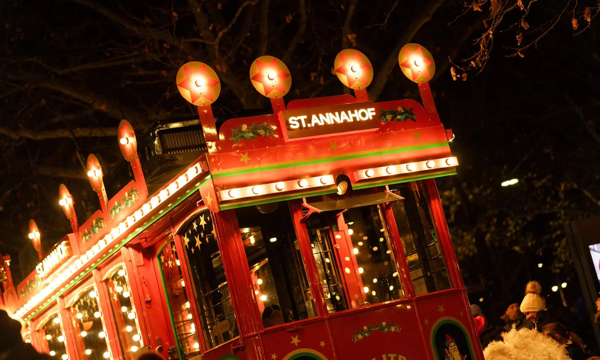 Beleuchtete rote Straßenbahn bei Nacht mit St. Annahof-Schild und Weihnachtsdekoration, darunter Menschen, die sich darunter versammeln.