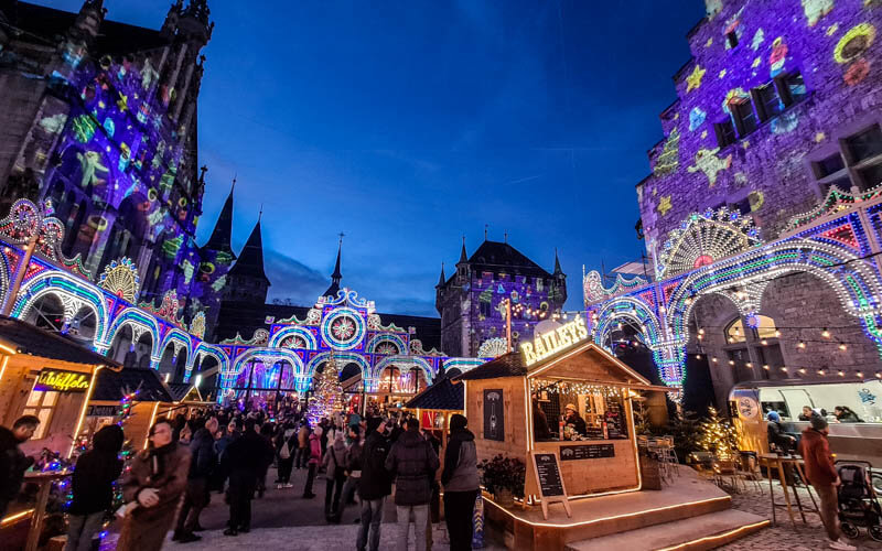 Belebter Weihnachtsmarkt bei Abenddämmerung mit festlich beleuchteten, historischen Gebäuden und Ständen, darunter ein beleuchtetes Holzhaus mit dem Schild "Barnes's". Die Fassaden sind mit bunten Weihnachtsprojektionen und Lichtern dekoriert, und Menschen schlendern durch die festliche Szenerie.