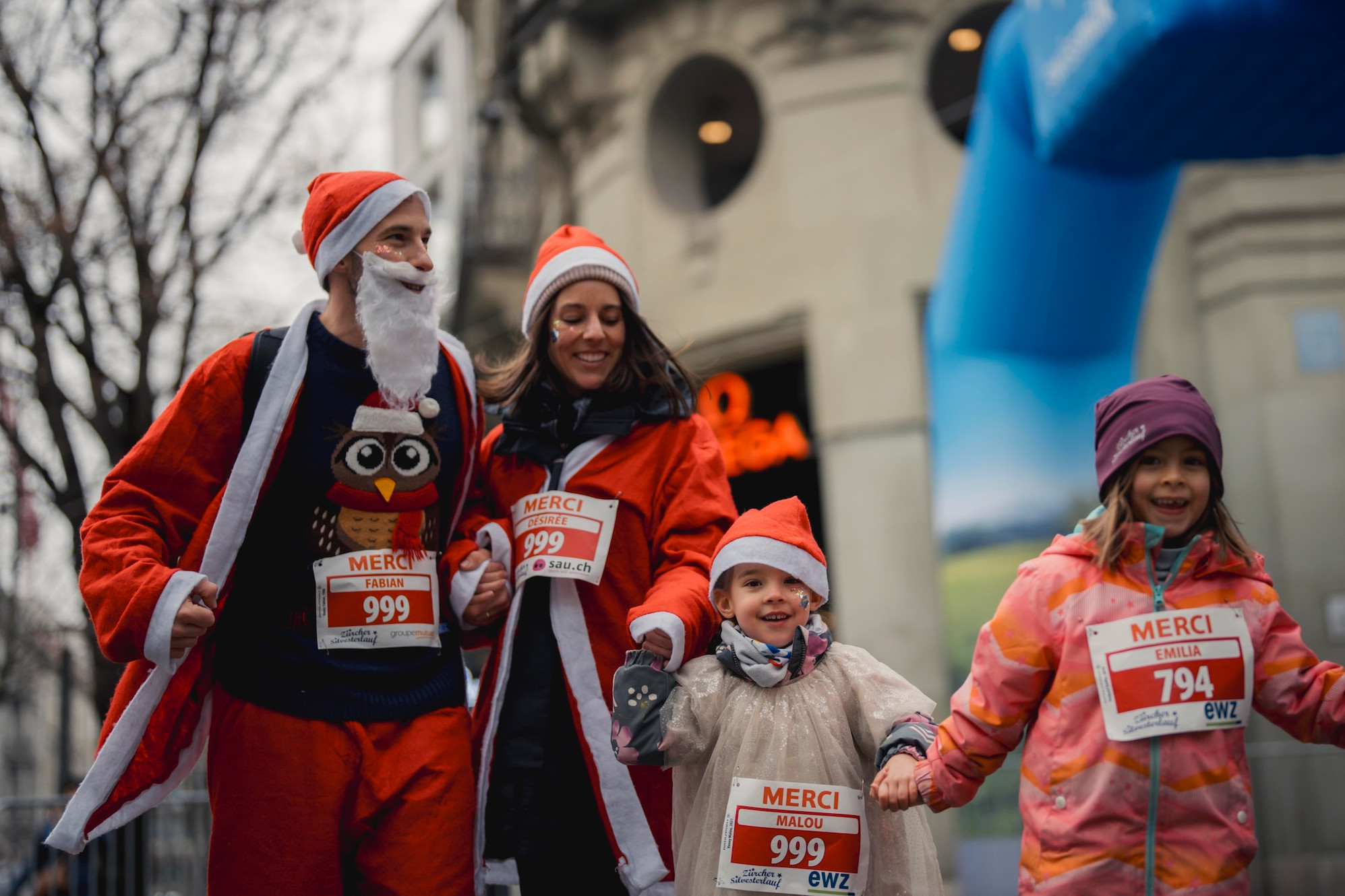 Vier fröhliche Menschen, zwei Erwachsene und zwei Kinder, in Weihnachtskostümen und mit Startnummern, die an einer Outdoor-Veranstaltung teilnehmen, halten sich an den Händen und lächeln.