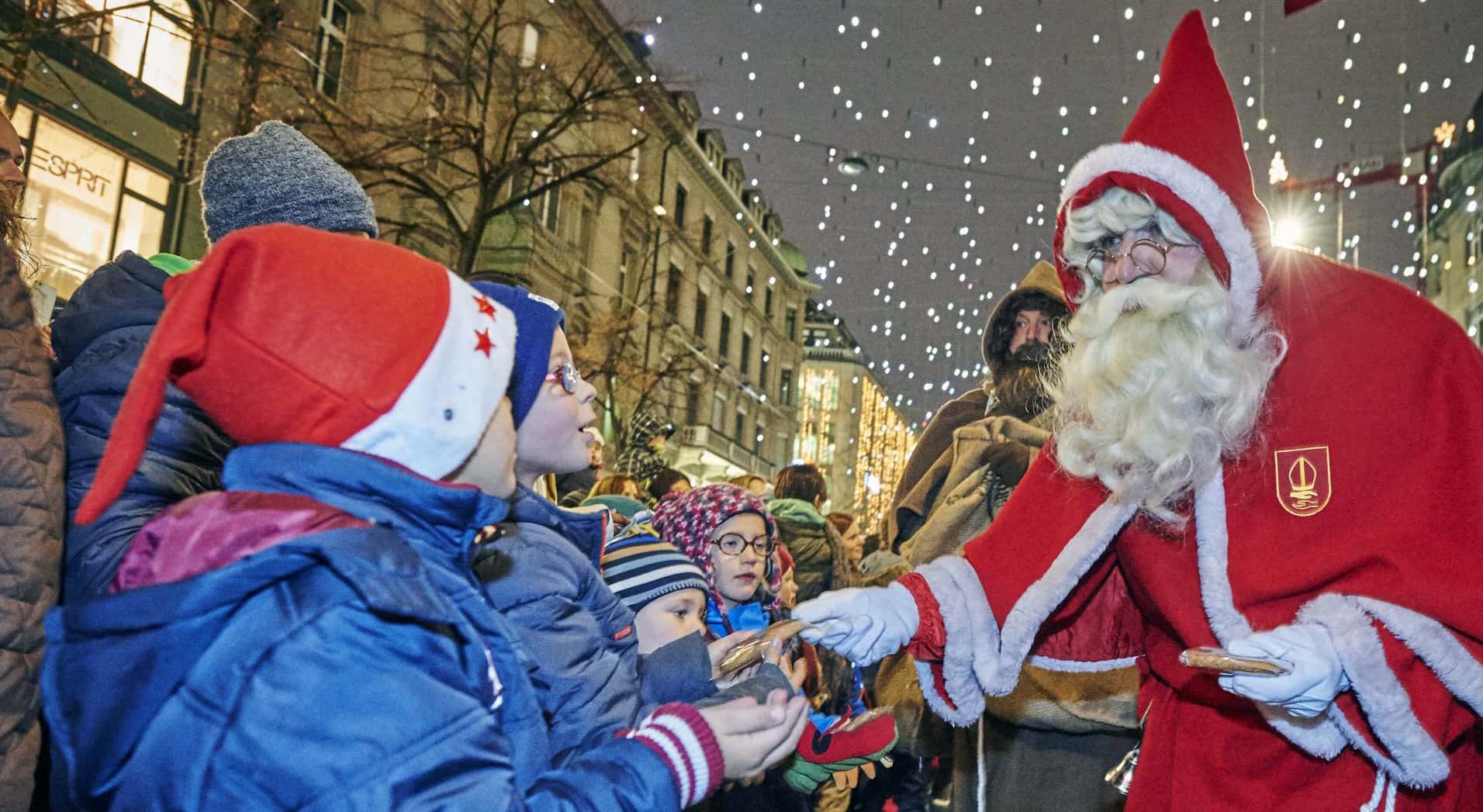 Samichlaus in rotem Mantel mit weissem Bart verteilt Gebäck an Kinder in warmen Winterjacken und Weihnachtsmützen auf beleuchteter Strasse bei einem Fest.