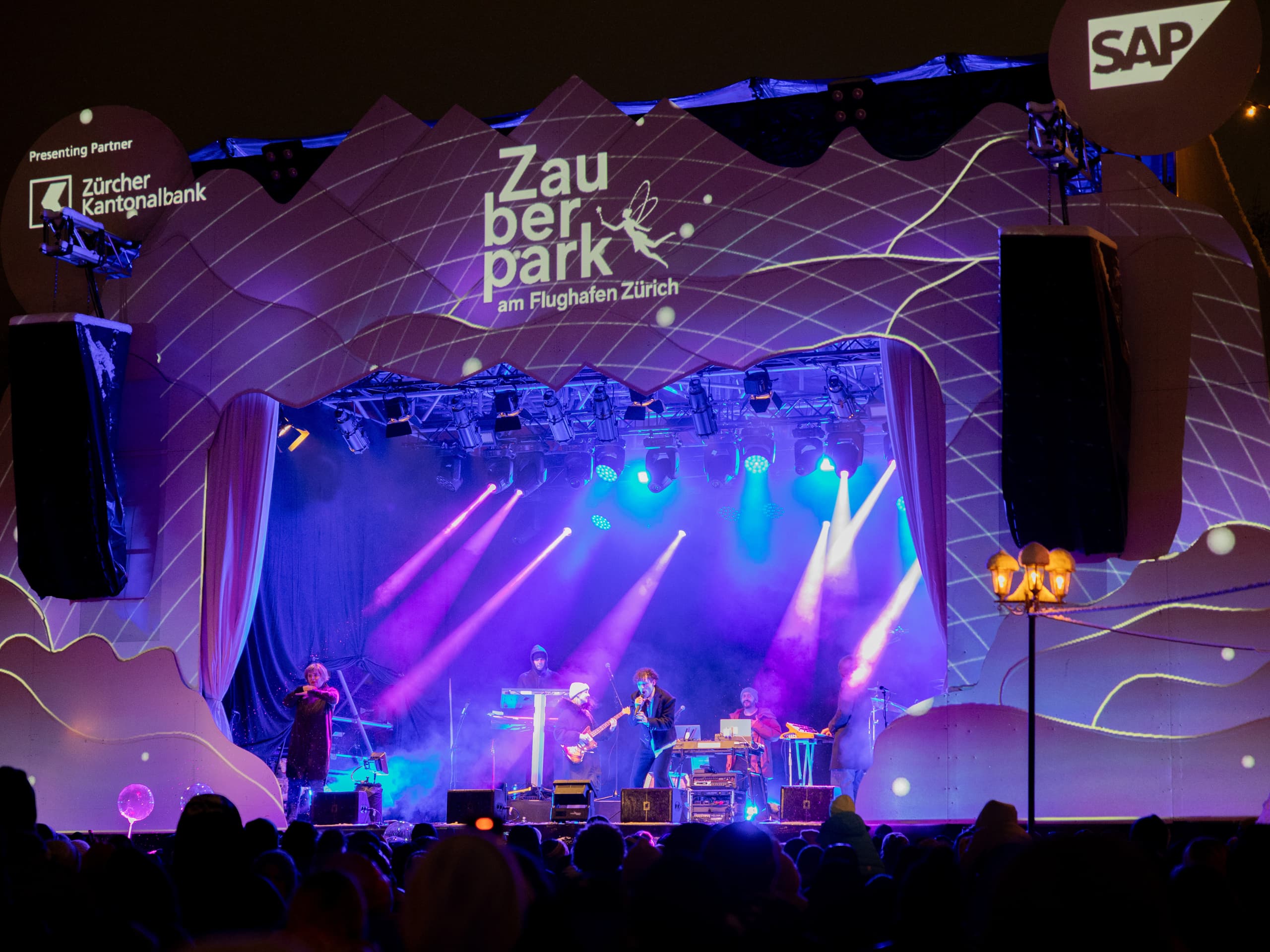Bühne mit Live-Bandperformanz bei Nacht, beleuchtet mit lila und blauen Scheinwerfern, Publikum im Vordergrund, Veranstaltungsname 'Zauberpark am Flughafen Zürich' auf Bühnenkulisse projiziert.