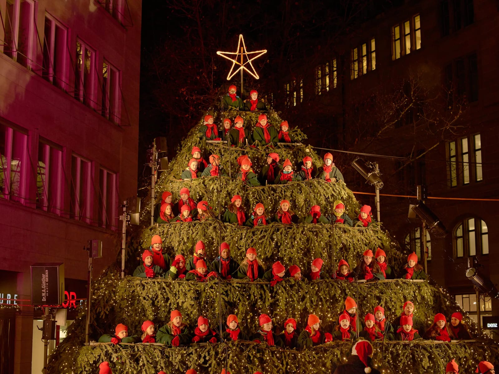 Kinder in roten Mützen und Schals singen auf einer großen, mit Tannenzweigen und Lichterketten kunstvoll gestalteten Pyramide mit einem leuchtenden Stern oben, vor einem dunklen Hintergrund aus beleuchteten Gebäuden in einer Stadt bei Nacht.