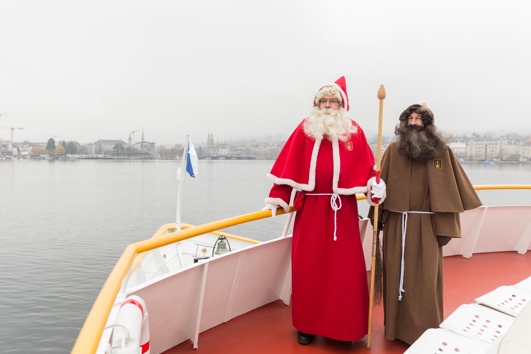 Zwei Männer in traditionellen Weihnachtskostümen auf dem Deck eines Schiffes, einer in einem roten Gewand mit weißem Pelzbesatz und roter Mütze, der andere in braunem Kapuzenumhang mit langem Bart; im Hintergrund ein nebliger See und eine Stadt mit Gebäuden und Kränen.