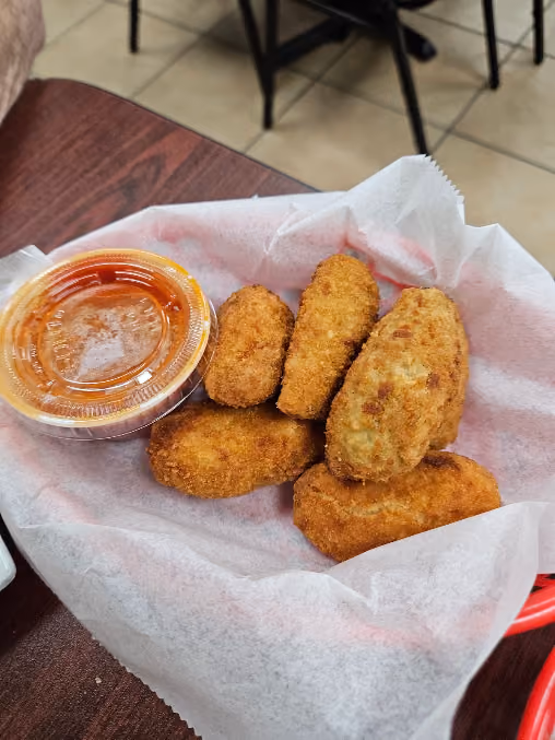 Basket lined with white paper holding five pieces of golden-brown fried jalapeño poppers next to a small container of orange dipping sauce on a wooden table.