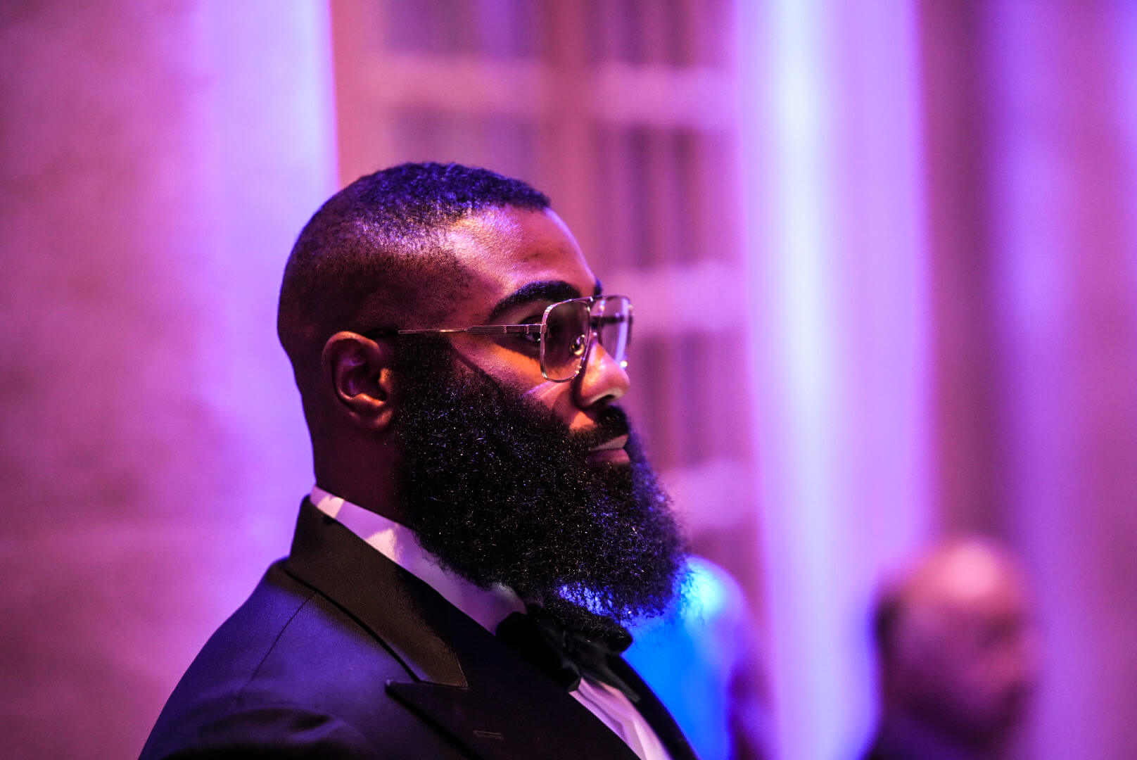 A guest in a tuxedo and glasses watches the program, lit in violet.