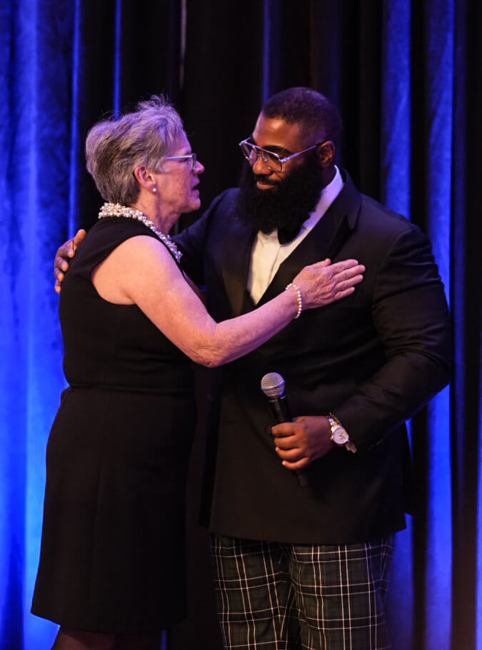 A woman and a man holding a microphone share an embrace on stage.
