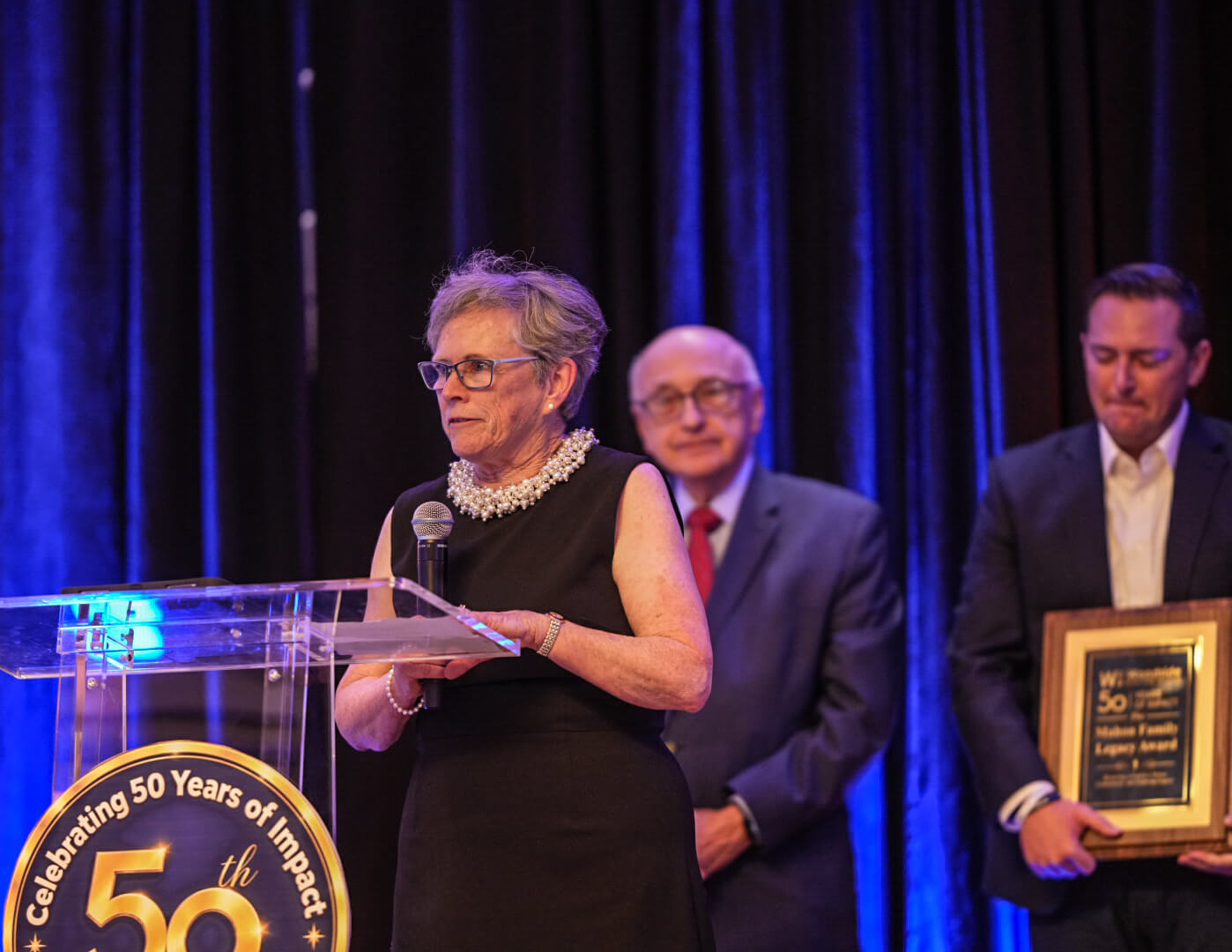 A speaker addresses the room from the 50th anniversary podium as two men wait beside her, one holding a framed award.