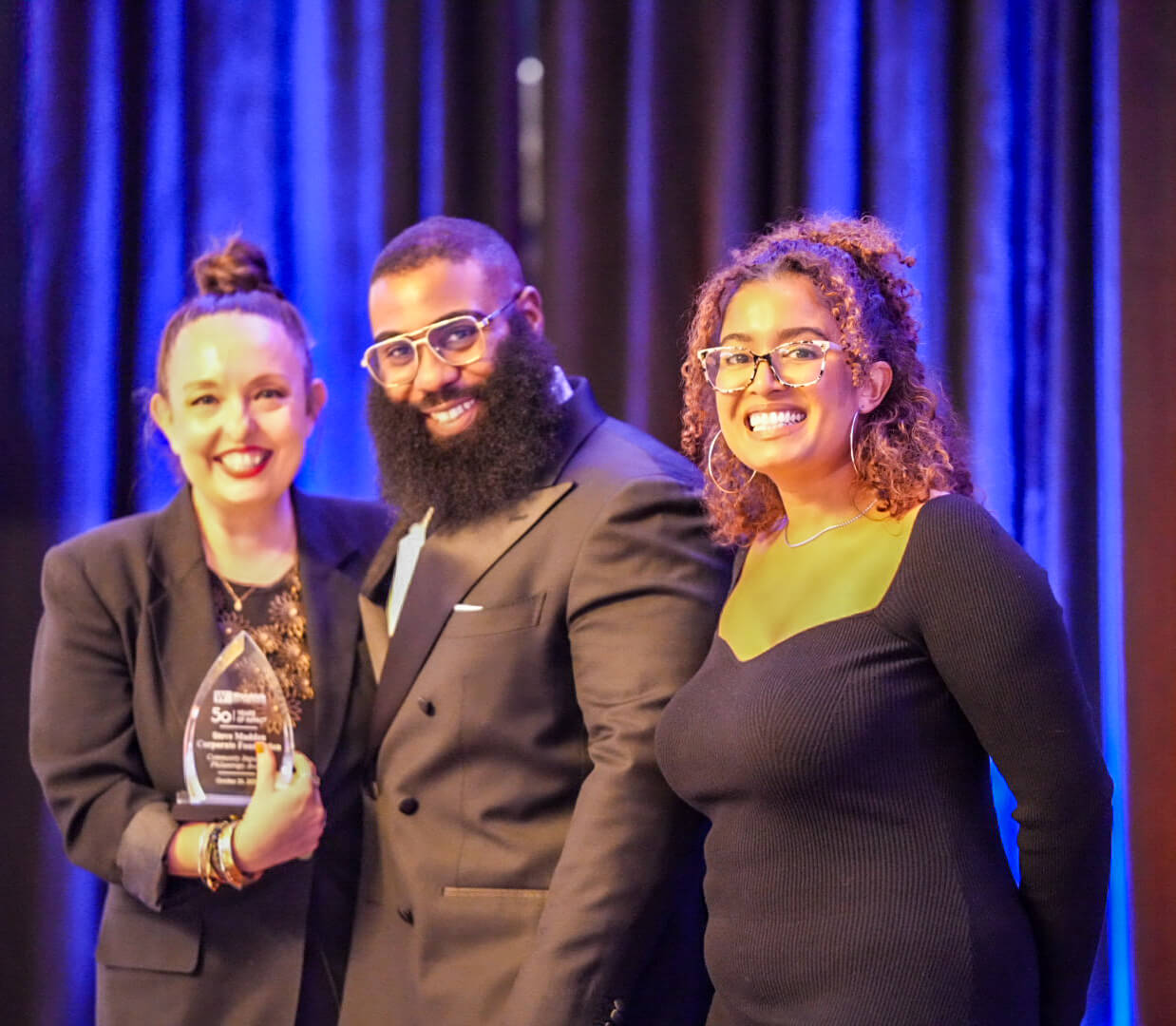 Three people smile on stage, one holding a crystal award.