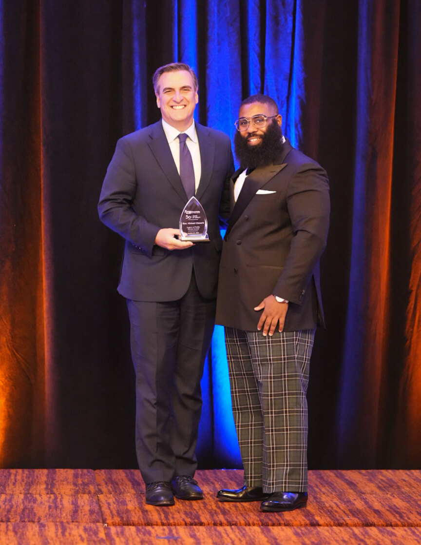Two men pose on stage with a crystal award in front of blue curtains.