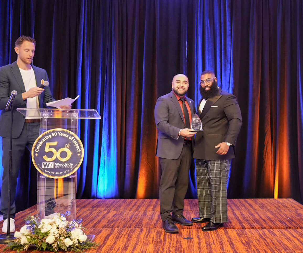 Two men stand on stage with an award as a presenter speaks at the 50th anniversary podium.