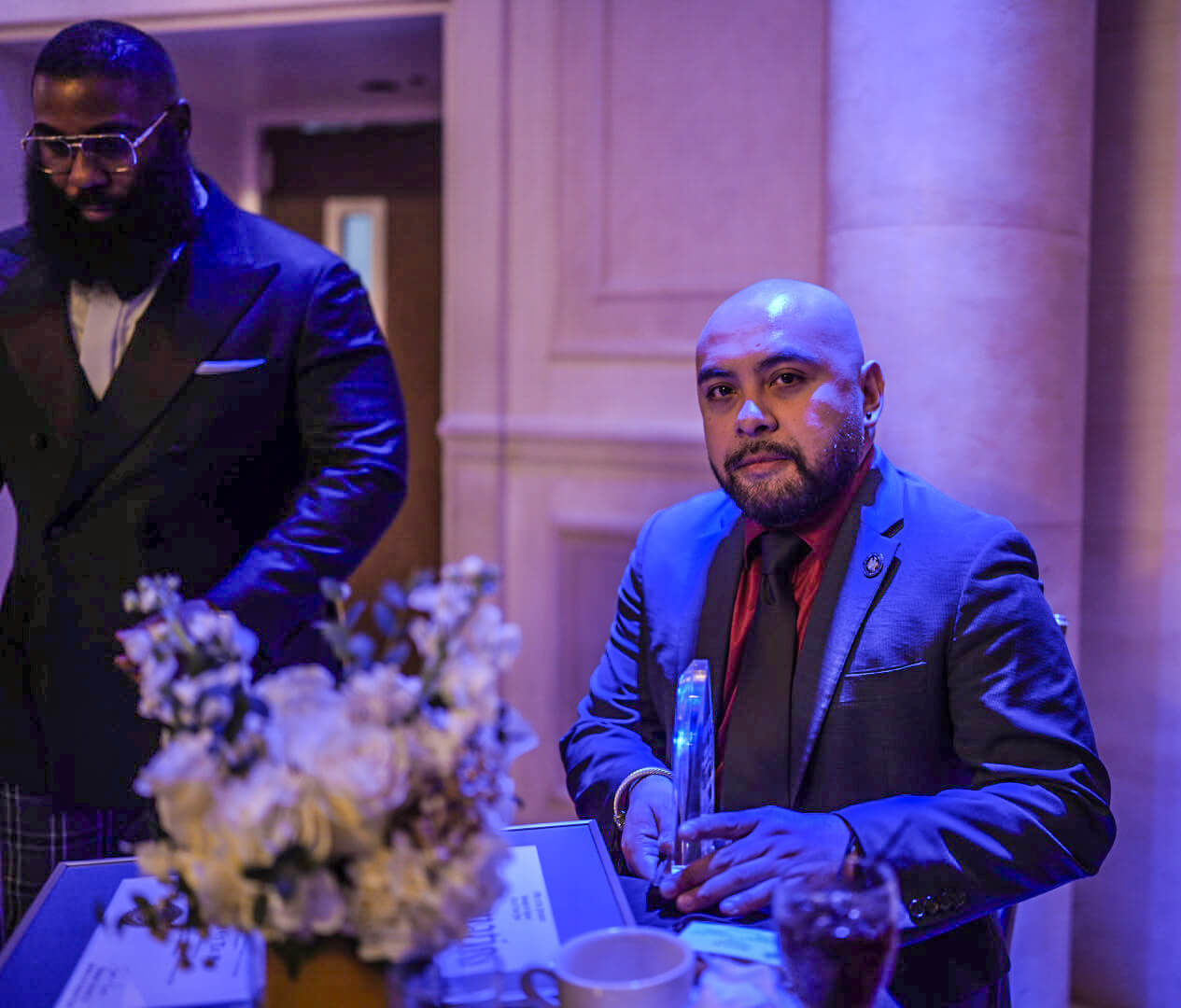 A guest seated beside a white floral centerpiece looks toward the stage under purple lighting.