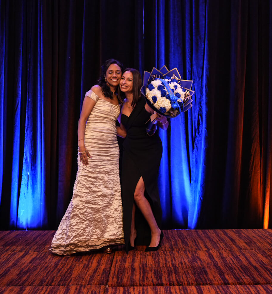 Two women pose on stage with a bouquet of blue and white flowers.