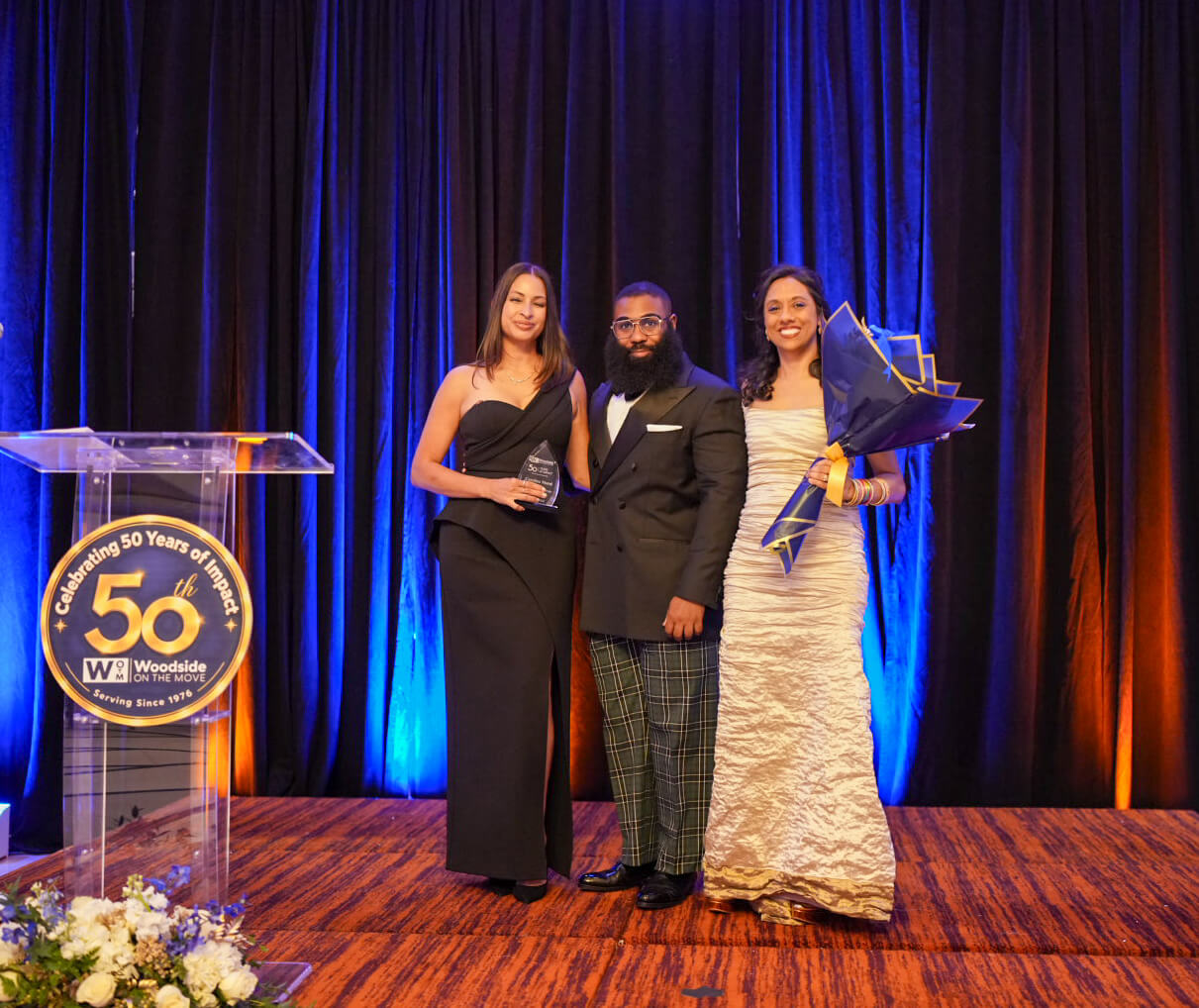 Three people pose beside the 50th anniversary podium, one holding an award and another a bouquet.