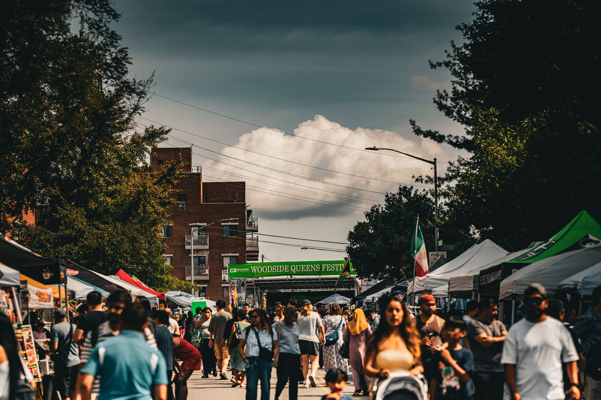 Crowded outdoor street festival with various vendor tents and people walking, under a partly cloudy sky, with a green banner reading 'Woodside Queens Fest'.