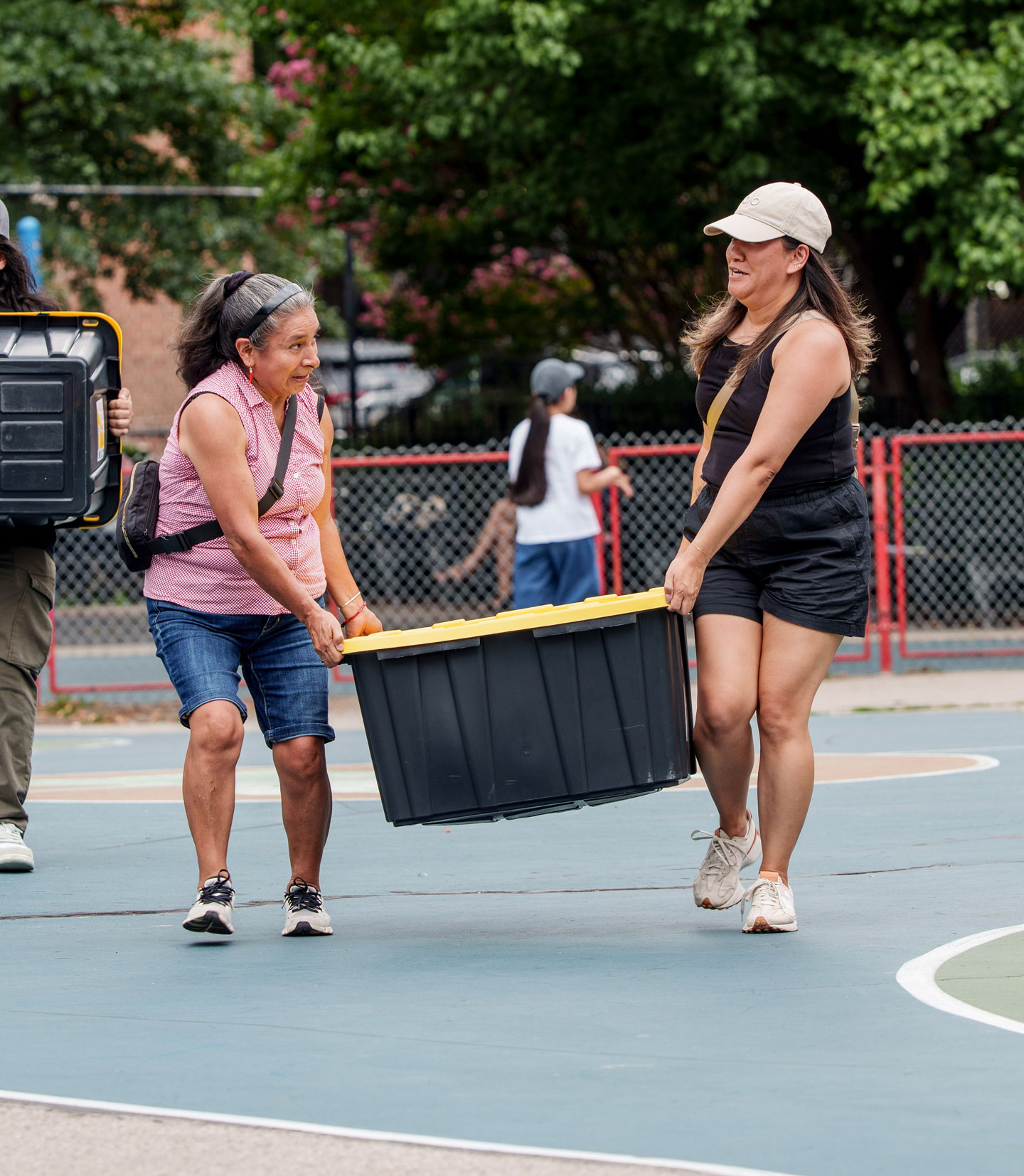 Two women outdoors carrying a large black plastic bin with a yellow lid together on a paved basketball court, with a chain-link fence and trees in the background.