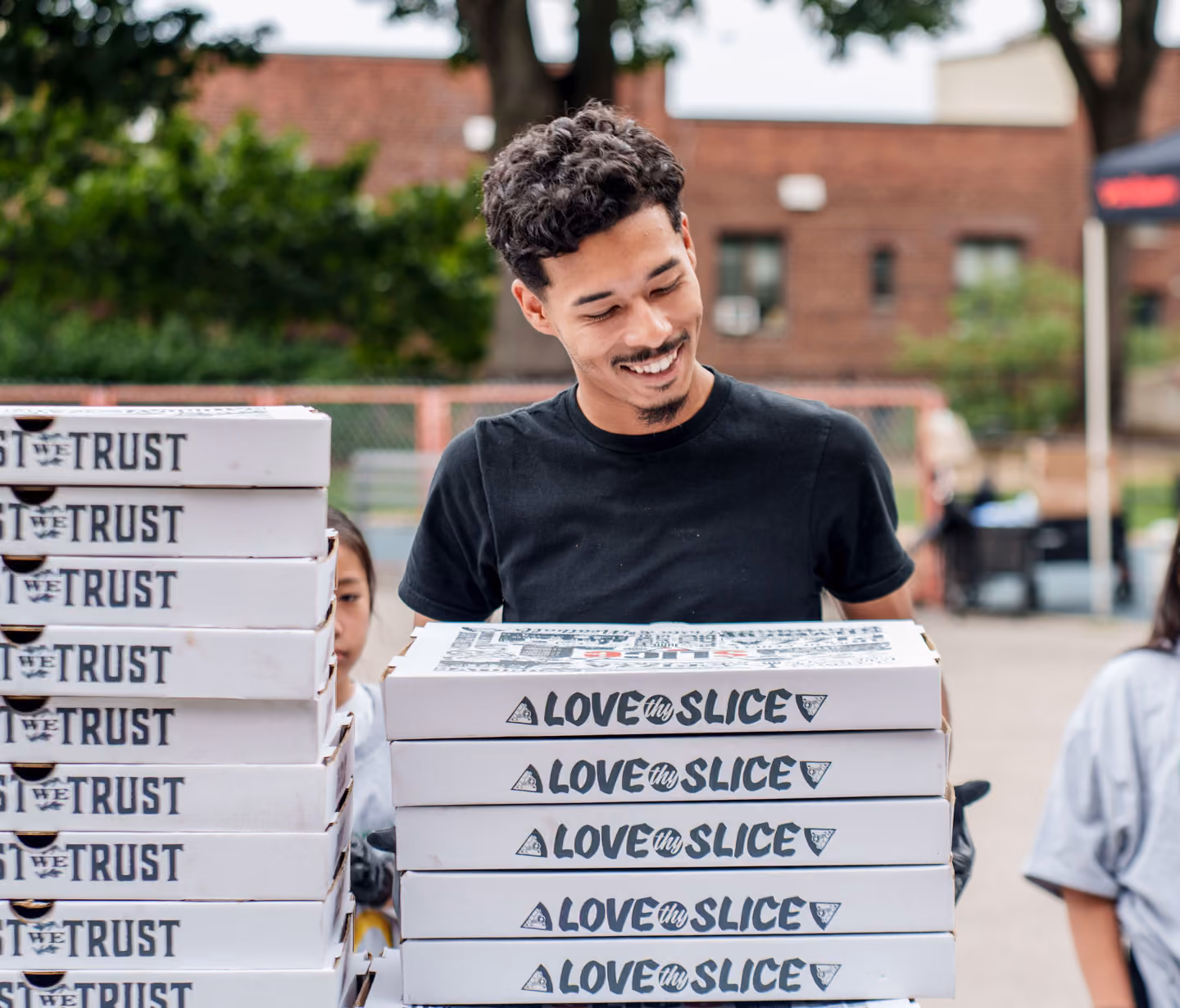 A smiling young man in a black shirt holding a stack of pizza boxes labeled 'LOVE my SLICE' outdoors with another stack of boxes labeled 'IN PIZZA WE TRUST' next to him.