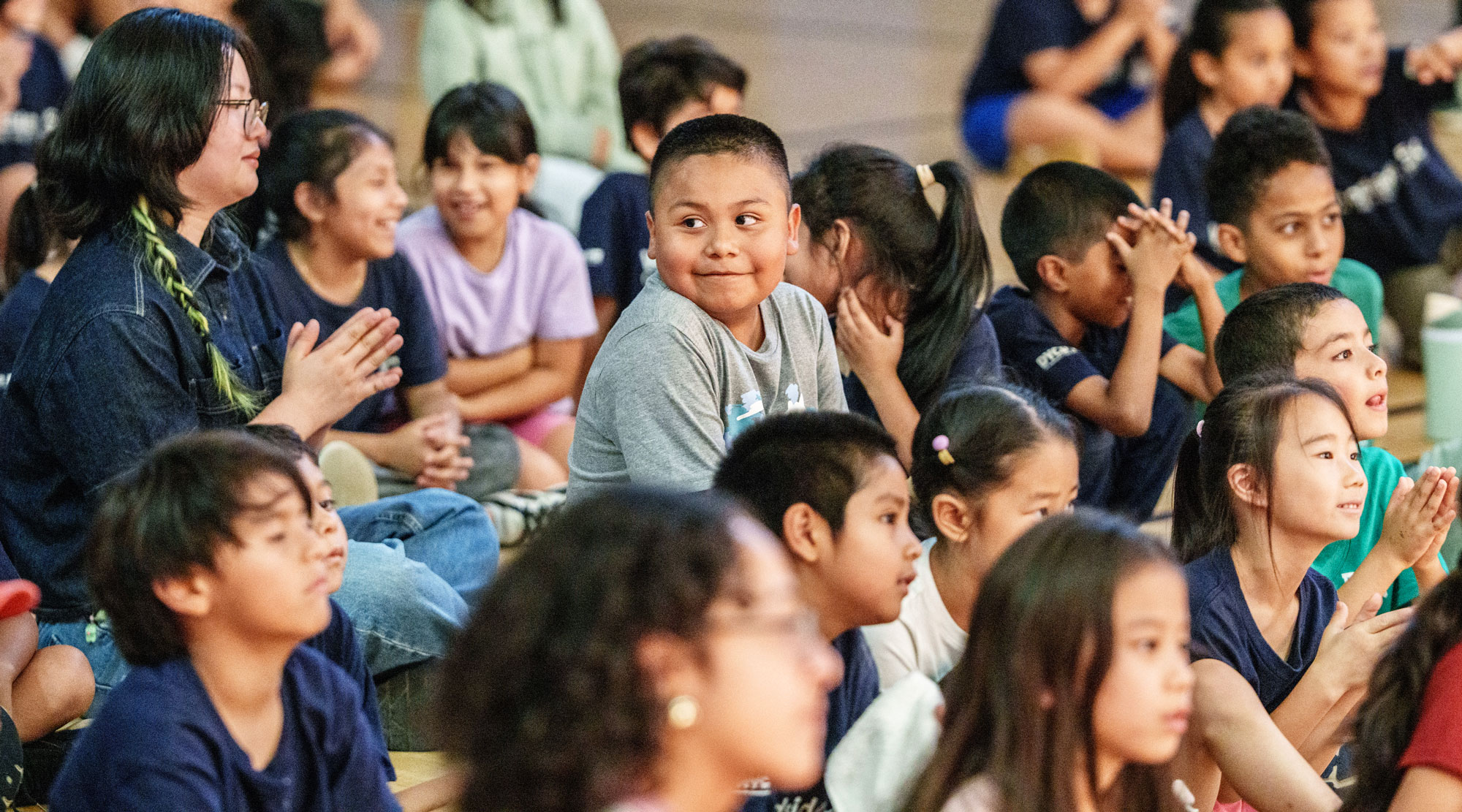 A diverse group of children sitting closely together indoors, some clapping and smiling, while an adult woman with glasses and a green braid also claps among them.