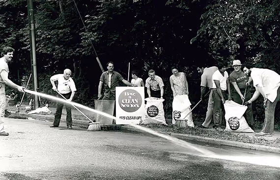 Black-and-white photo of six people cleaning a street with brooms and holding 'I love a CLEAN New York' signs during the 1979 cleanup.