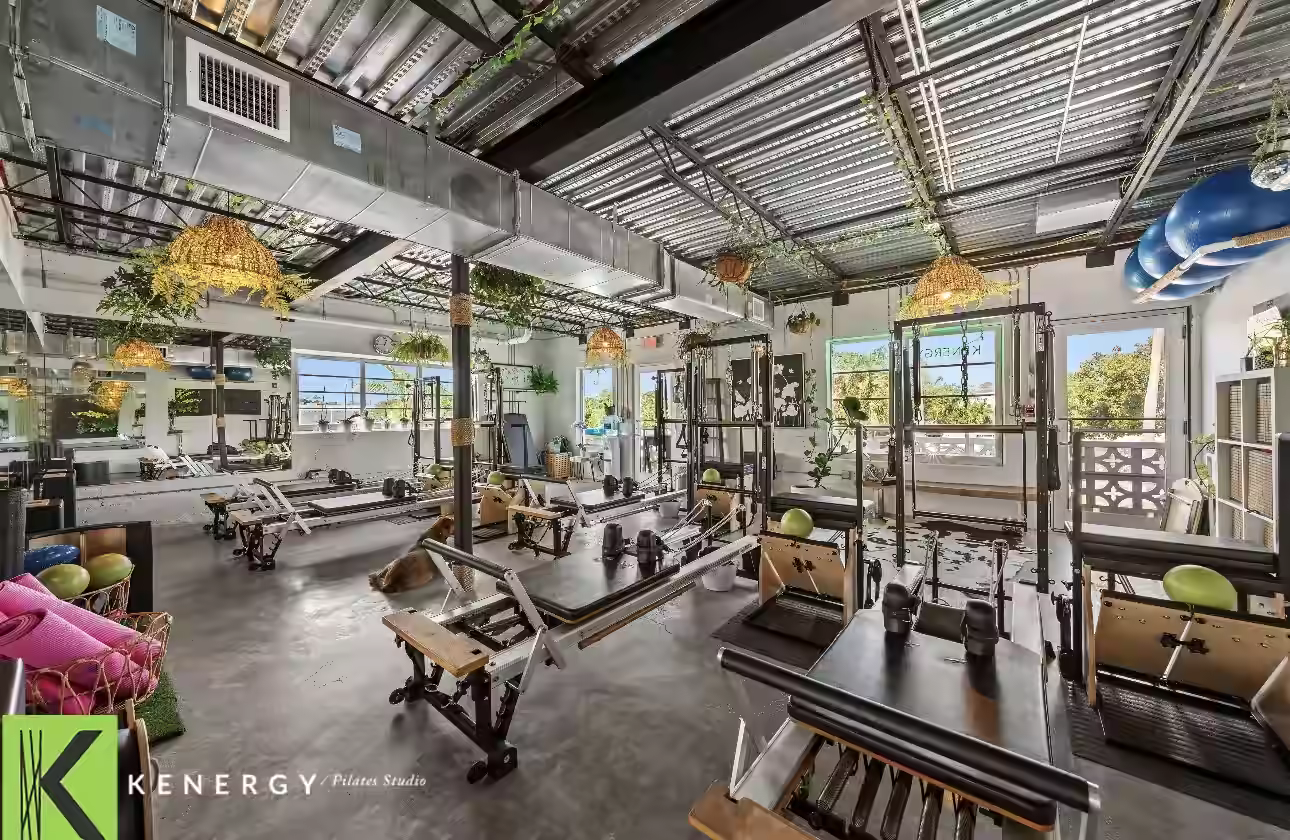 Pilates studio with reformers, hanging plants, mirrors, and large windows.