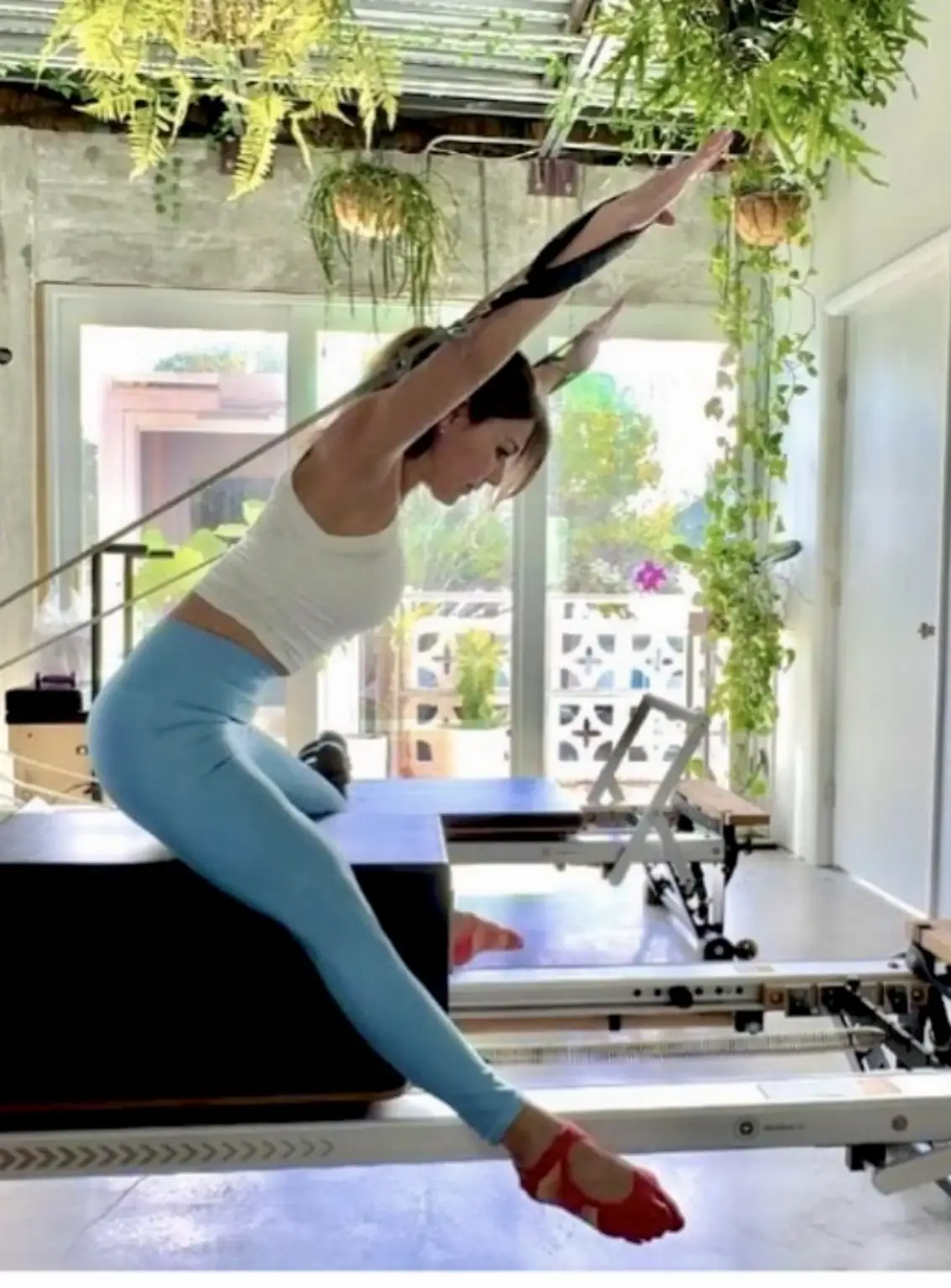 Person in a white top and blue leggings exercising on a Pilates reformer with arms extended overhead in resistance straps.