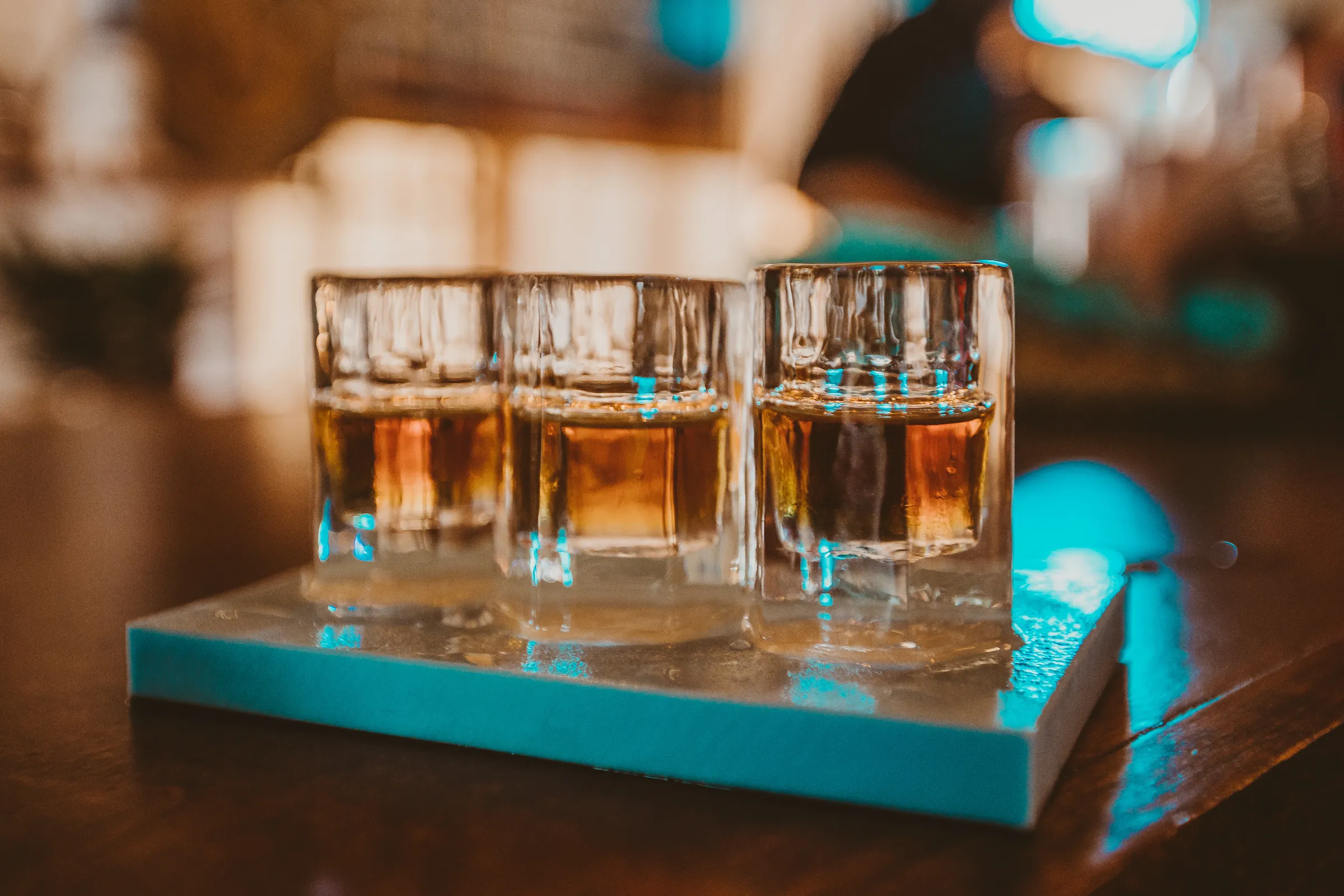 Three glasses of bourbon on a teal tasting board, set on a wooden bar with warm lights glowing in the background.