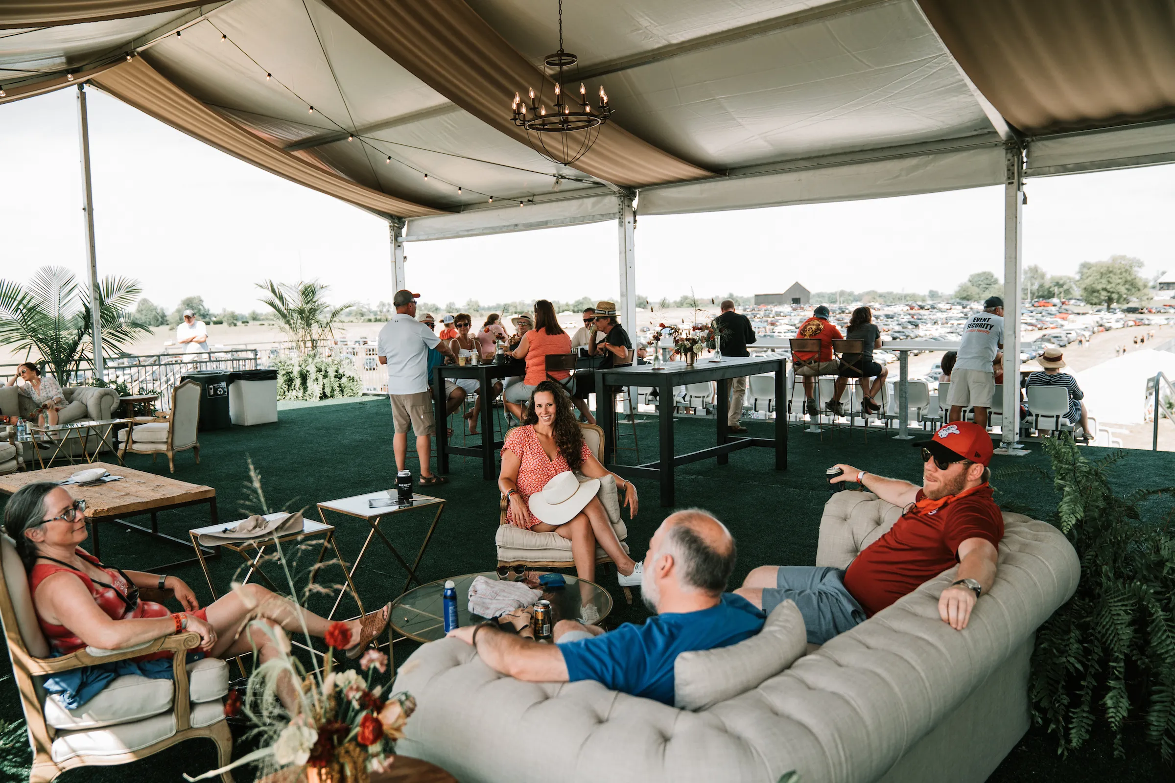 Guests relaxing on tufted sofas and armchairs in a shaded lounge tent with a chandelier, overlooking the festival grounds.