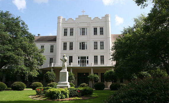 Jesuit Spirituality Center at St. Charles College, located in Grand Coteau, Louisiana. A white multi-story building with castle-like battlements and a cross on top, surrounded by green trees and bushes, with a statue of a person holding a book and a child in the foreground on a grassy area.
