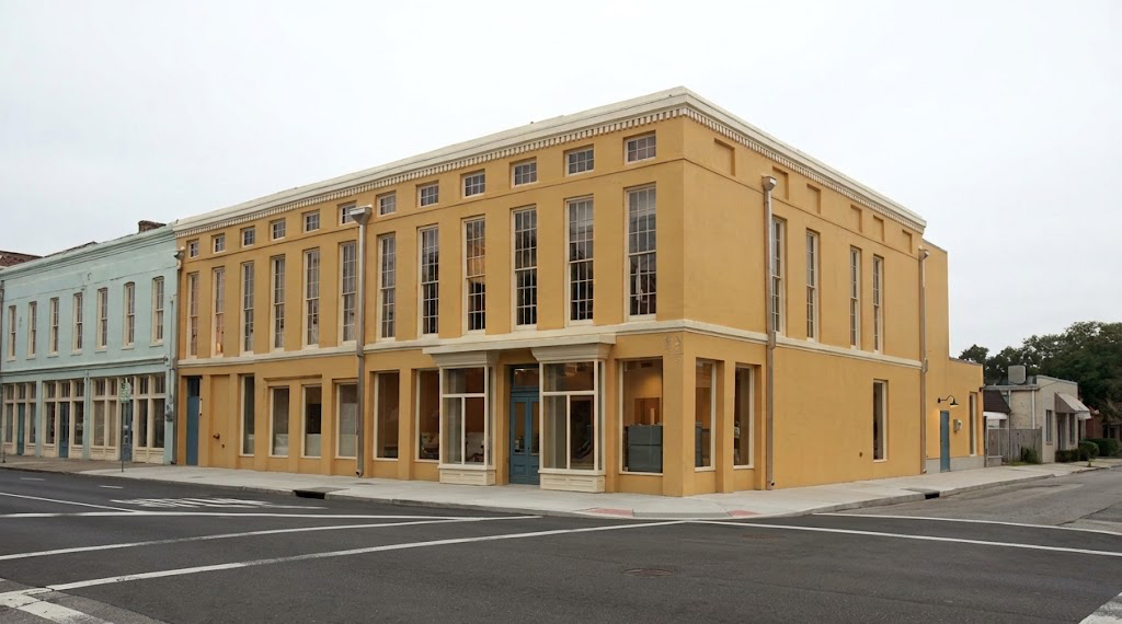 Corner view of a two-story building with a yellow facade and tall, narrow windows, located on an empty street intersection under a cloudy sky.