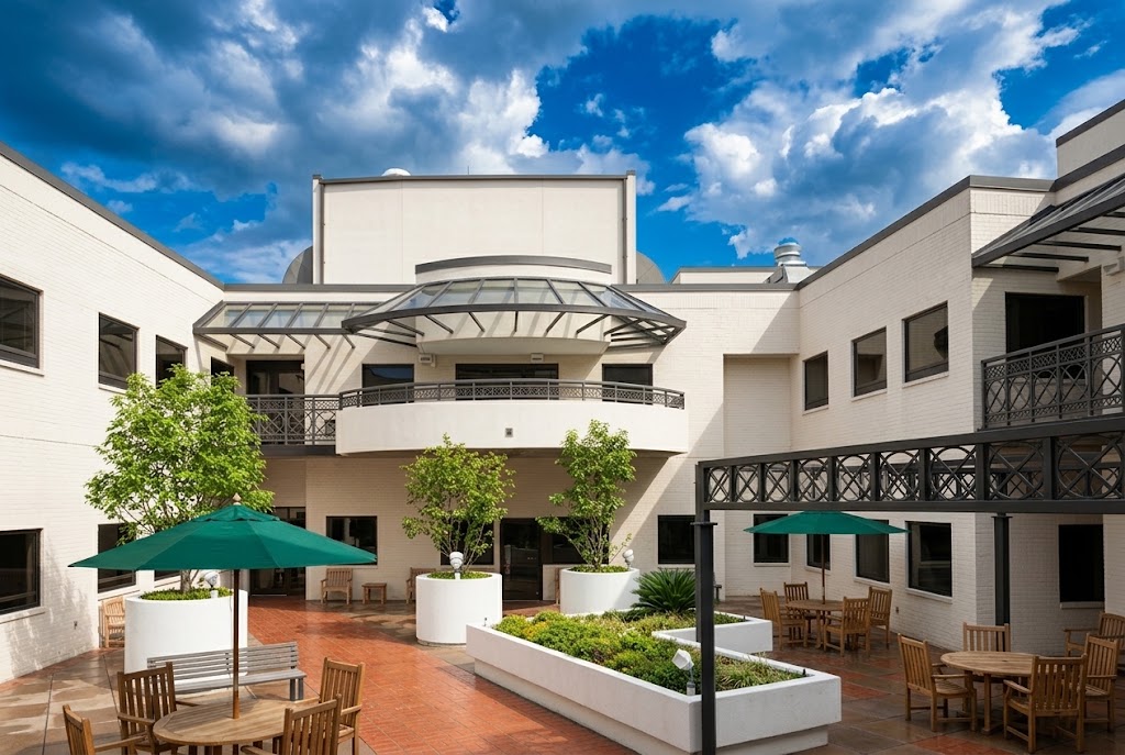 Outdoor courtyard with white buildings surrounding a tiled floor, green umbrellas shading wooden tables and chairs, large potted trees, and a brick red walkway under a partly cloudy blue sky.