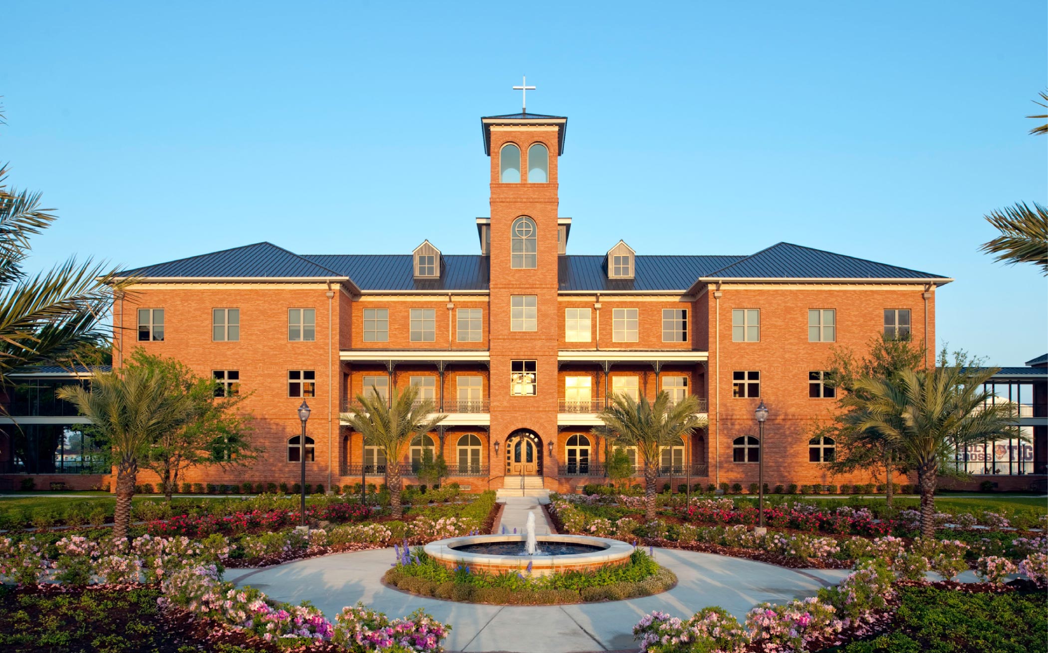 Holy Cross High School - New Orleans, LA. A large brick building with a central tower topped by a white cross, surrounded by landscaped gardens with palm trees and flowering plants under a clear blue sky.