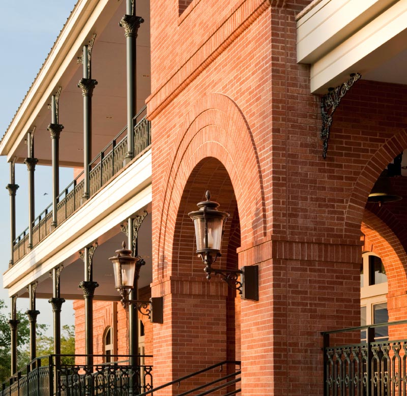 Two-story brick building facade with large arched openings, black ornate iron railings and columns, and vintage-style wall lanterns mounted on brick pillars.