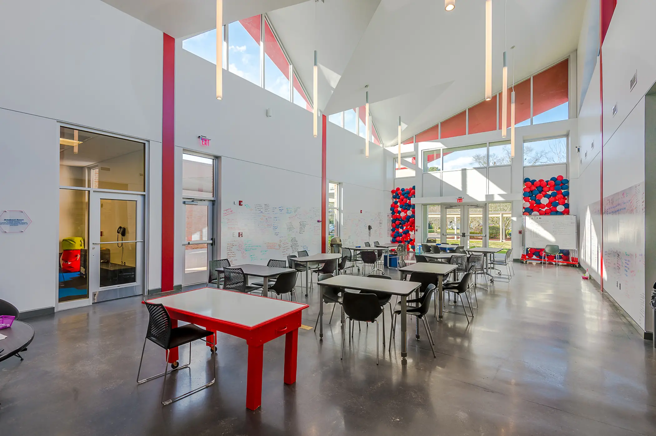 Bright, spacious classroom with multiple rectangular tables and black chairs on polished concrete floor, white walls featuring red accent stripes, high angular ceiling with hanging cylindrical lights, large windows letting in natural light, whiteboards with writing on walls, and red, white, and blue balloon decorations near the entrance doors.