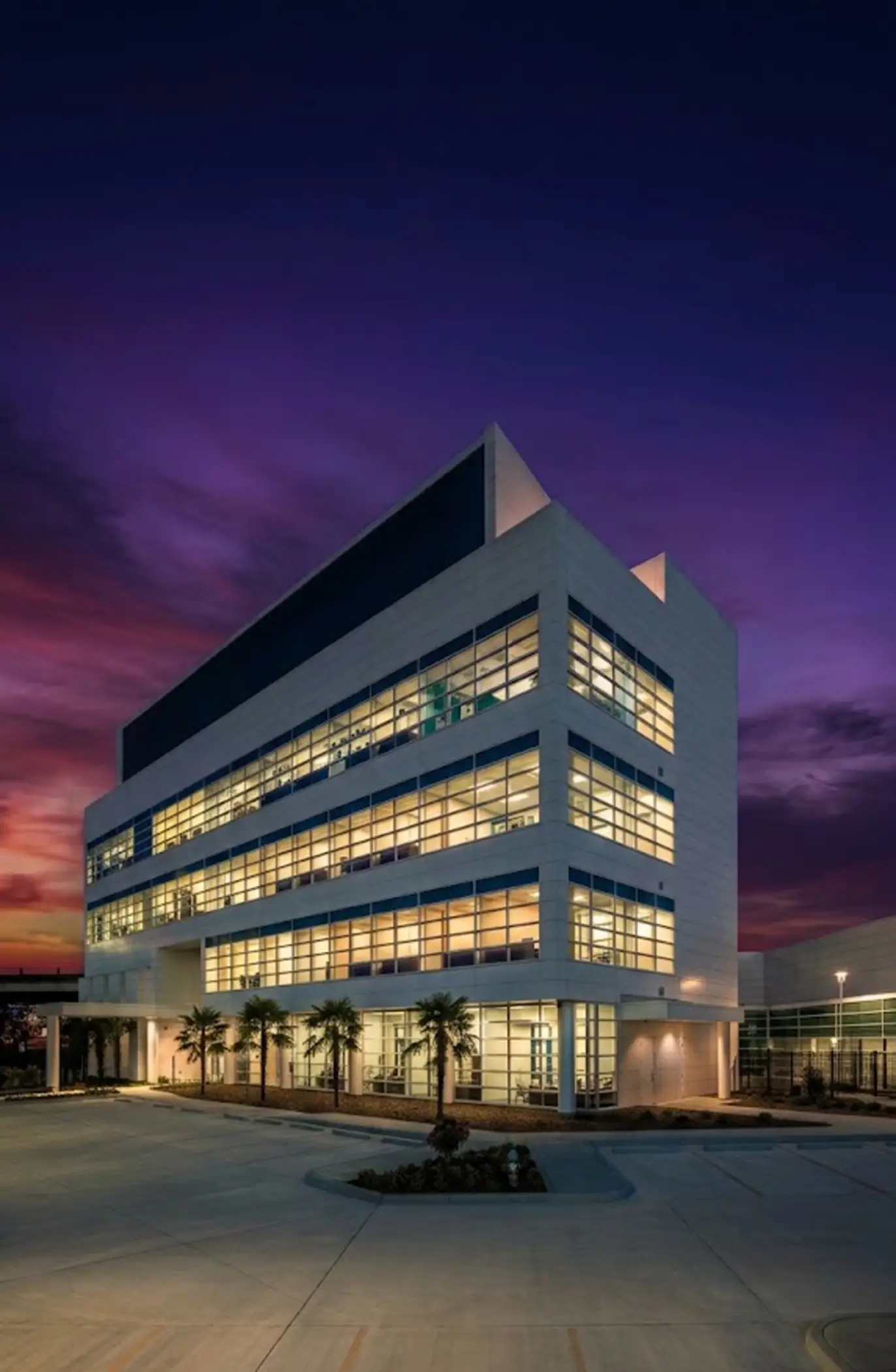 Modern multi-story office building with large illuminated windows at dusk, surrounded by palm trees and an empty parking lot.