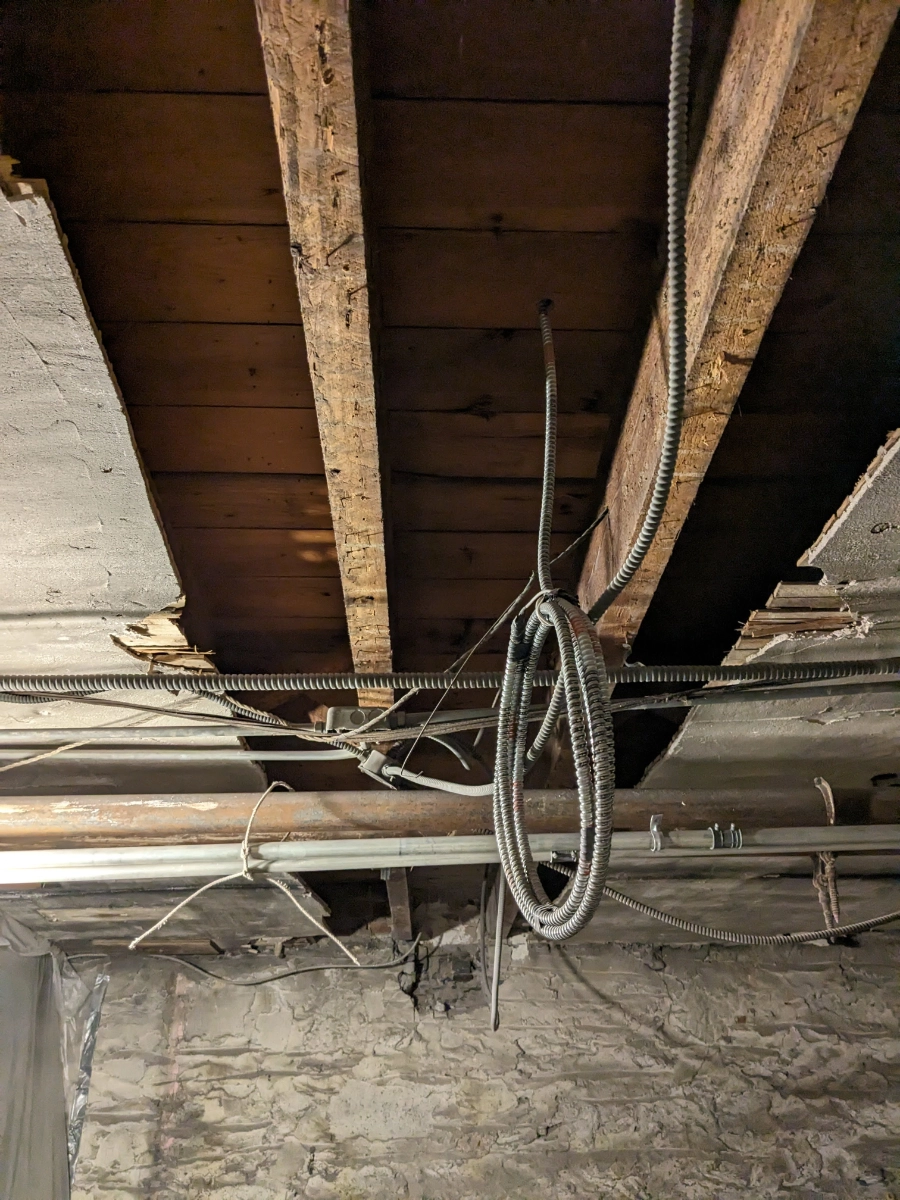 Exposed ceiling with wooden beams and various pipes and wires in an unfinished basement.