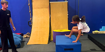 A young girl is jumping over a blue padded block in an indoor gym area, with a man watching her and two wooden ramps in the background.