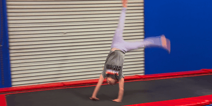 Child performing a handstand tumble on a trampoline with red edges in an indoor gym setting with blue walls and beige shutter backdrop.