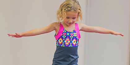 Young girl with blond curly hair wearing a colorful sleeveless top and dark shorts, balancing with arms outstretched against a plain gray background.