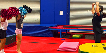 Three children practicing cheerleading indoors on a red mat with pom-poms raised, one child stands farther in black with arms raised without pom-poms.