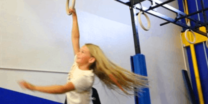 Blonde woman swinging from gymnastic rings indoors, with hair flowing and a joyful expression, in a gym setting with blue and yellow equipment.