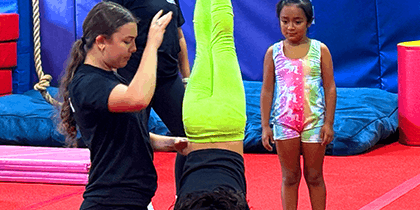 Gymnastics coach assisting a child performing a handstand while another child in a colorful leotard watches, on a red gym floor with blue mats.