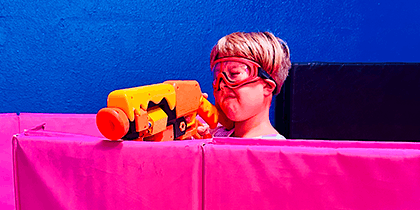 Boy wearing red goggles aiming a yellow and orange foam dart blaster over a pink barrier in an indoor play area with blue walls.