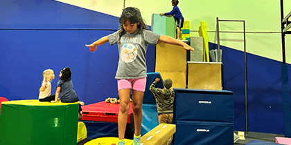 Children playing in an indoor gym with padded equipment; one girl balances on a beam while others climb or sit nearby.