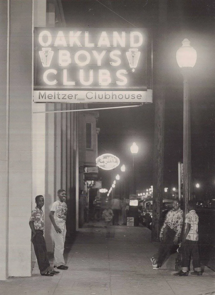 The lit "Oakland Boys Clubs" neon sign above the Meltzer Clubhouse entrance at night, with boys standing on the sidewalk (vintage photograph)
