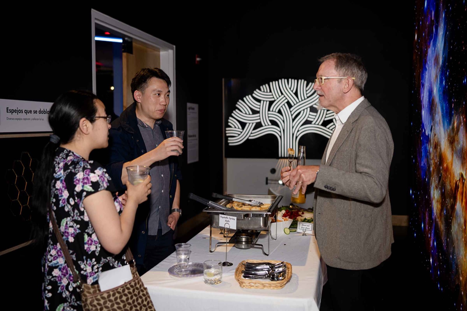 A gray-haired man in a blazer chats with two guests holding cups beside a buffet table at an evening reception, with the white Oakland tree logo on the wall behind them