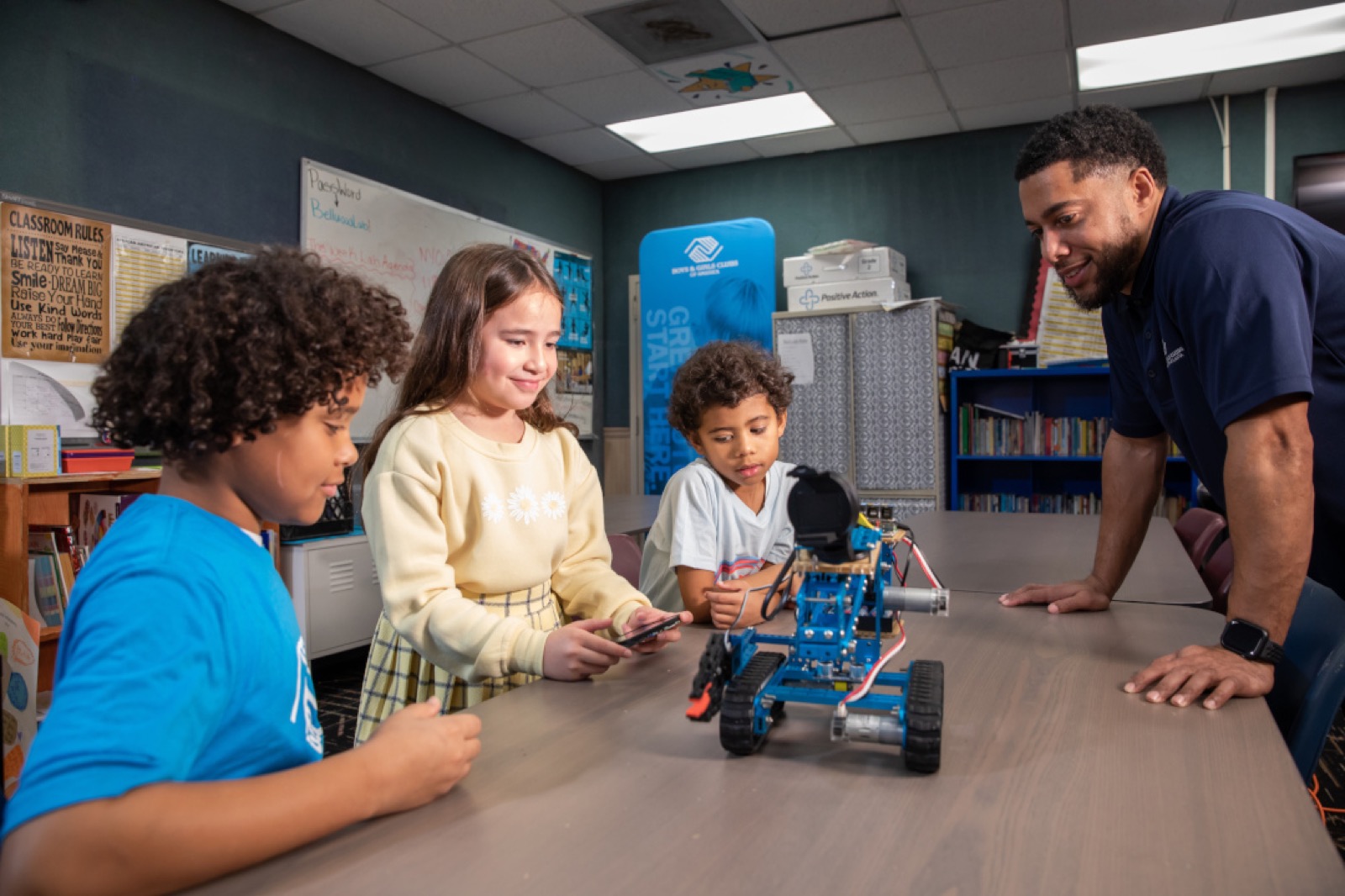 Three children and a Boys & Girls Clubs staff member gather around a small blue tracked robot on a table in a clubhouse classroom.